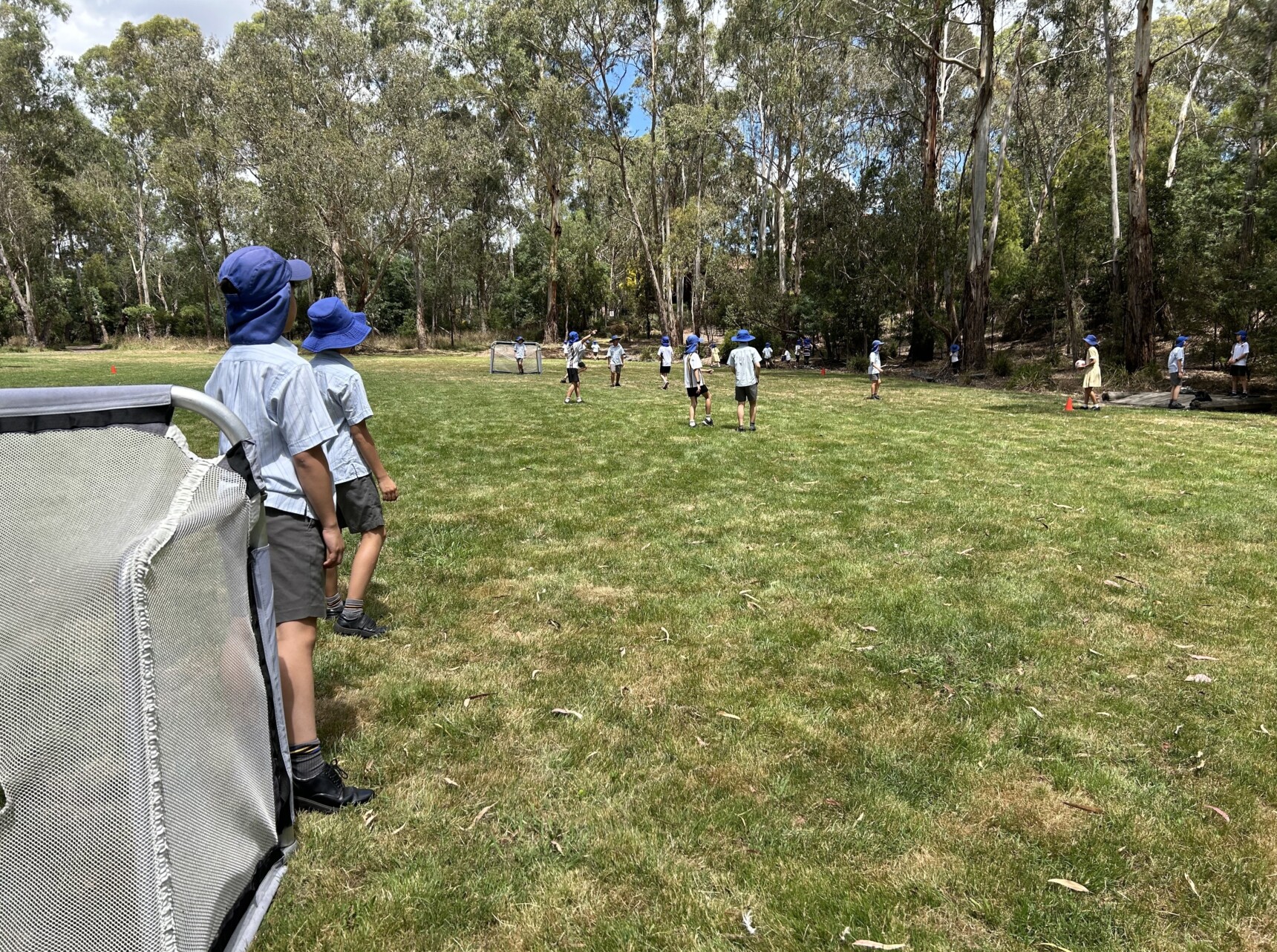Students from Donvale Private School participate in an outdoor sports activity on a grassy field surrounded by tall eucalyptus trees. Some children stand near a small soccer goal, wearing blue hats and school uniforms, while others engage in the game in the distance. The natural setting provides a picturesque backdrop for the school’s outdoor activities.