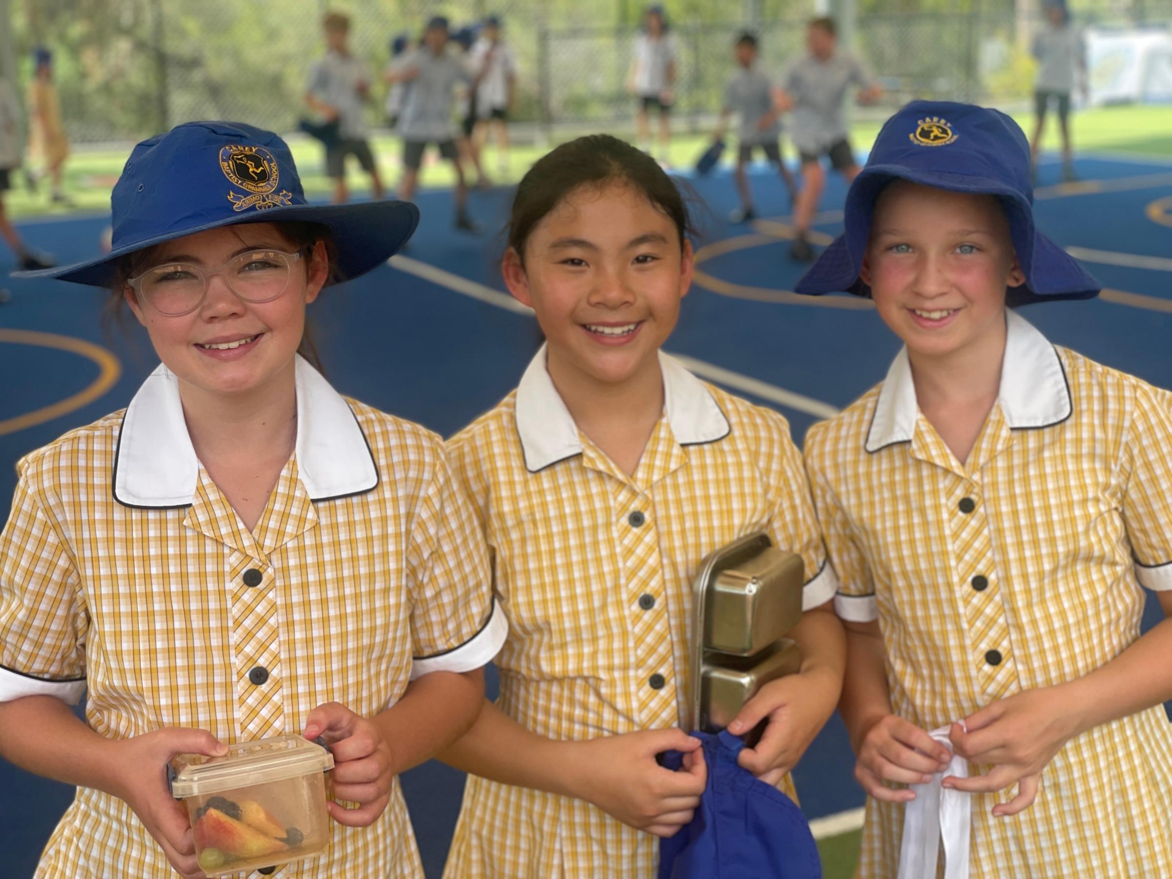Three smiling students from Donvale Private School stand together on an outdoor sports court, dressed in yellow checkered school uniforms with white collars. Two of them wear blue wide-brim hats featuring the school emblem. They hold lunch containers and bags, with a background of other students playing on the blue sports court. The girls appear happy and engaged in their school environment.