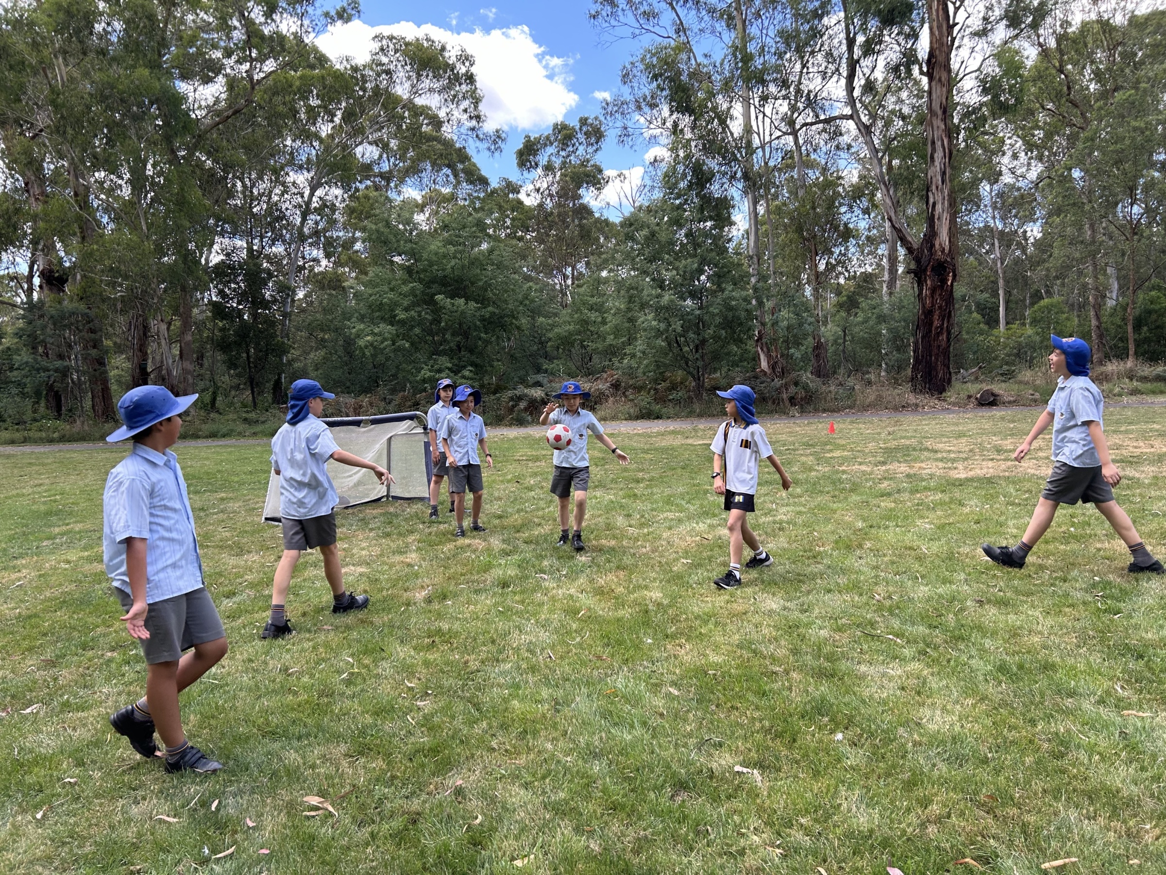 Students from Donvale Private School engage in a friendly soccer game on a grassy field surrounded by lush eucalyptus trees. Dressed in blue sun hats and school uniforms, the children actively participate, with one student holding a soccer ball while others move around the field. A small goalpost is visible in the background, adding to the lively outdoor sports scene.