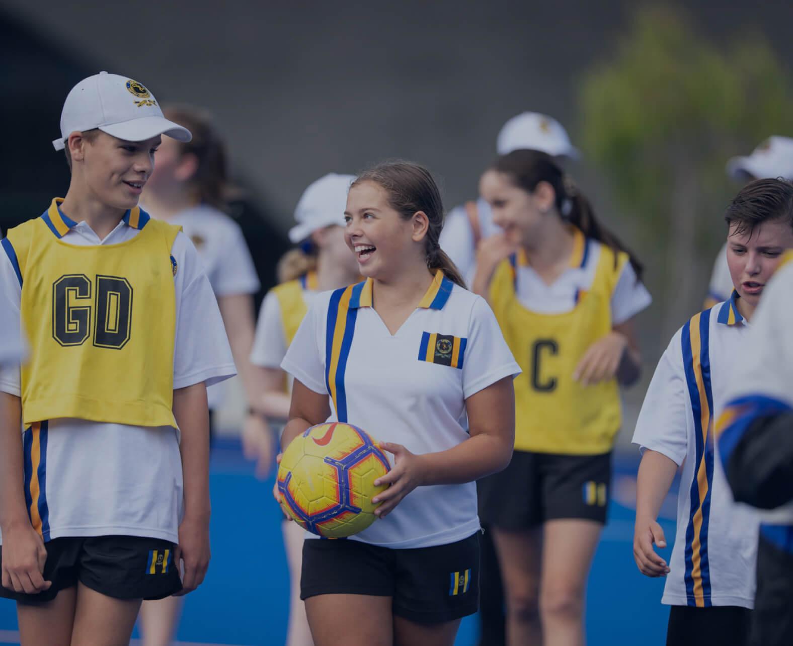 Students at Bulleen Sports Campus engage in an energetic outdoor sports game on a blue court. Wearing white sports uniforms with blue and gold accents, some players wear yellow bibs indicating their team positions. A smiling girl holds a yellow and blue soccer ball while chatting with a teammate wearing a 'GD' bib. The scene reflects teamwork, camaraderie, and active participation in school sports.