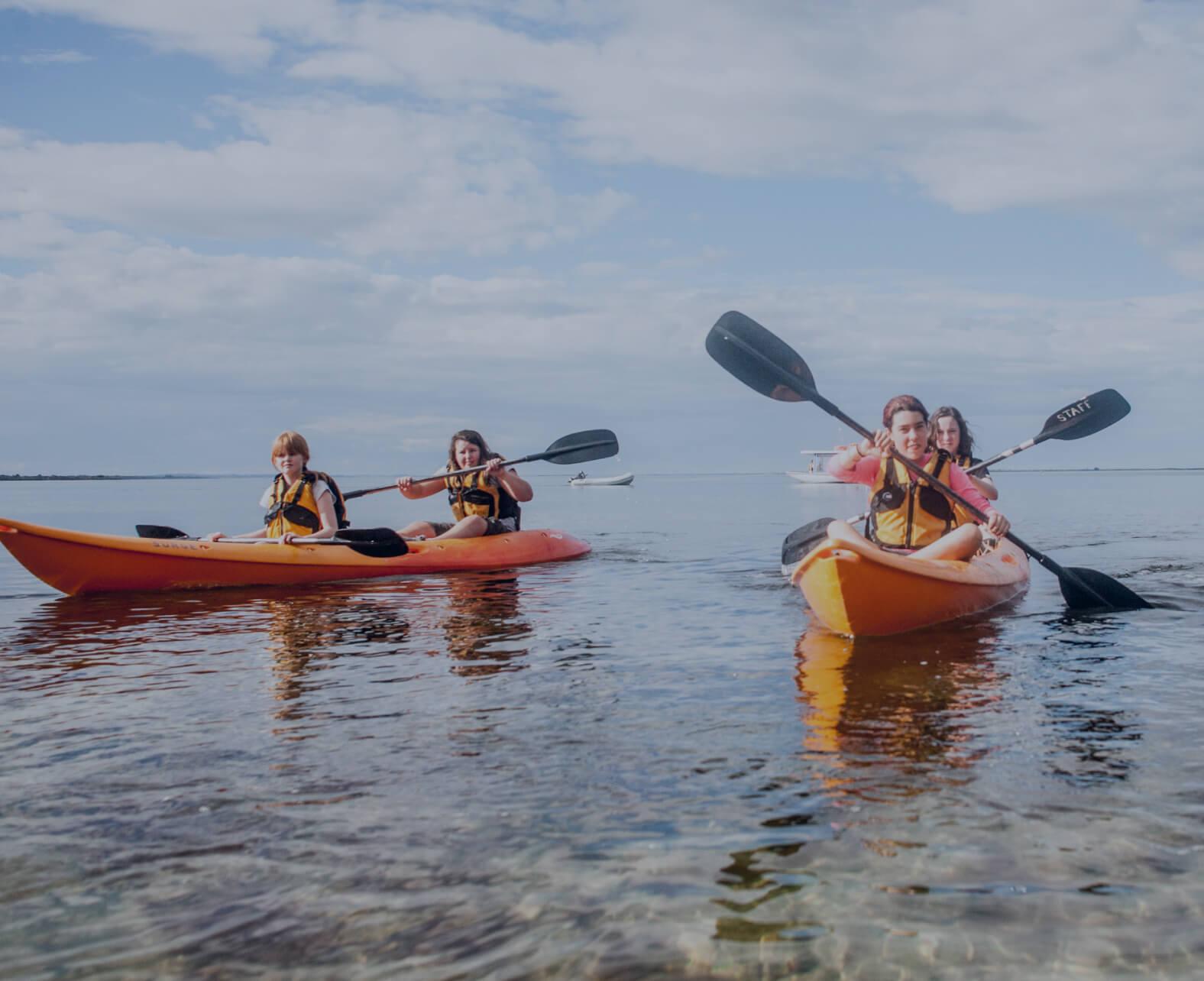 Students enjoy a kayaking activity at Camp Toonallook, paddling across calm, clear waters. Wearing life jackets, they navigate bright orange kayaks in pairs under a partly cloudy sky. The scenic coastal setting, with boats visible in the background, highlights an adventurous and immersive outdoor camp experience.