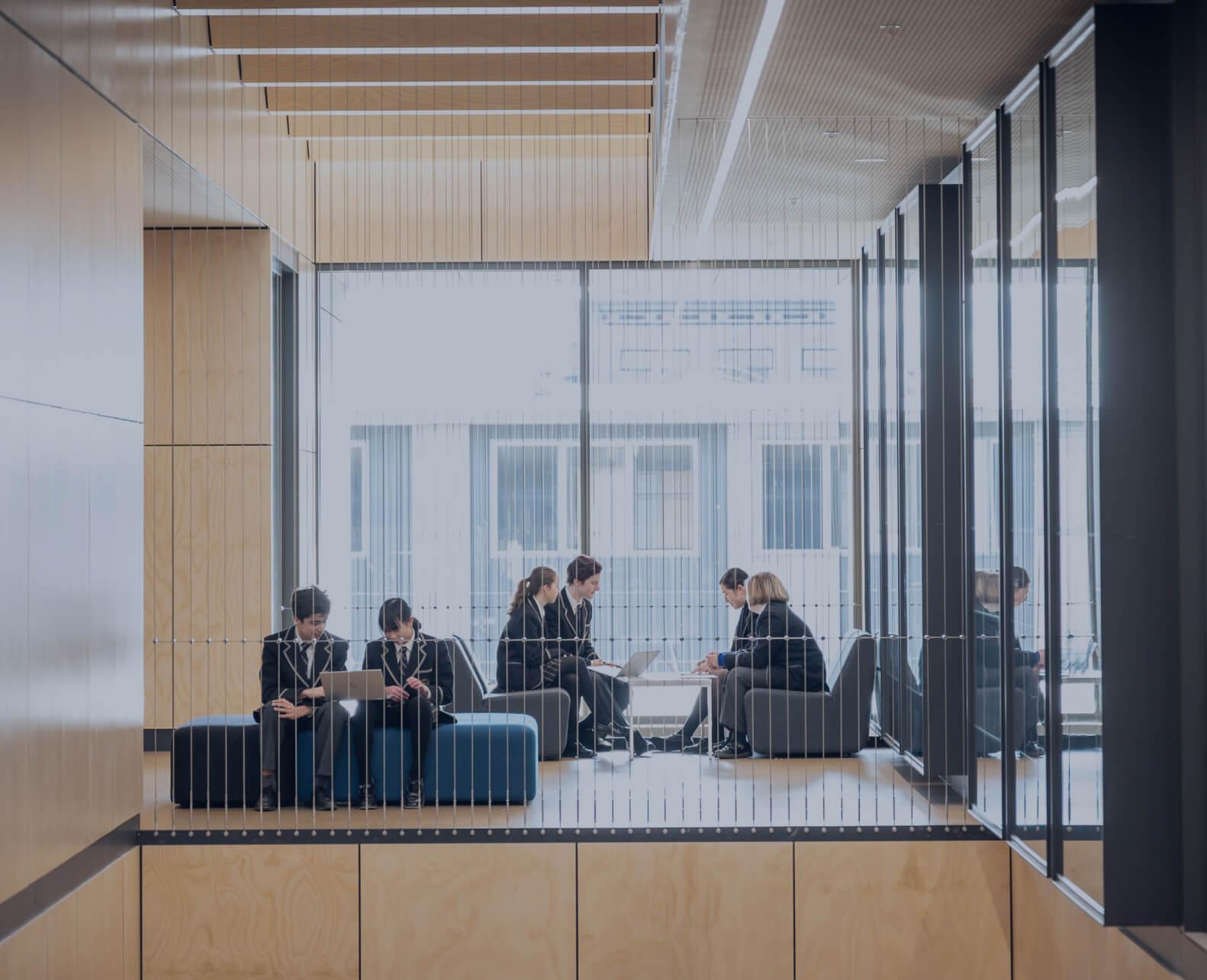 A group of six people are sitting in a modern, open space with large windows at Carey Kew Campus Private School, engaged in discussions and using laptops, surrounded by a minimalist wooden interior.