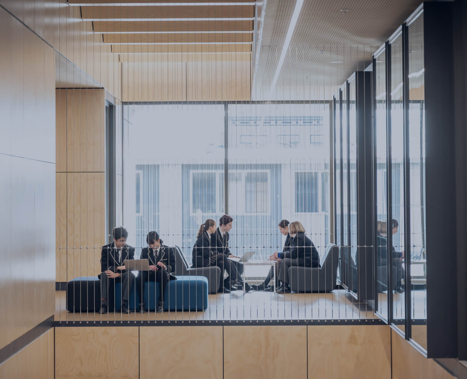 A group of six people are sitting in a modern, open space with large windows at Carey Kew Campus Private School, engaged in discussions and using laptops, surrounded by a minimalist wooden interior.